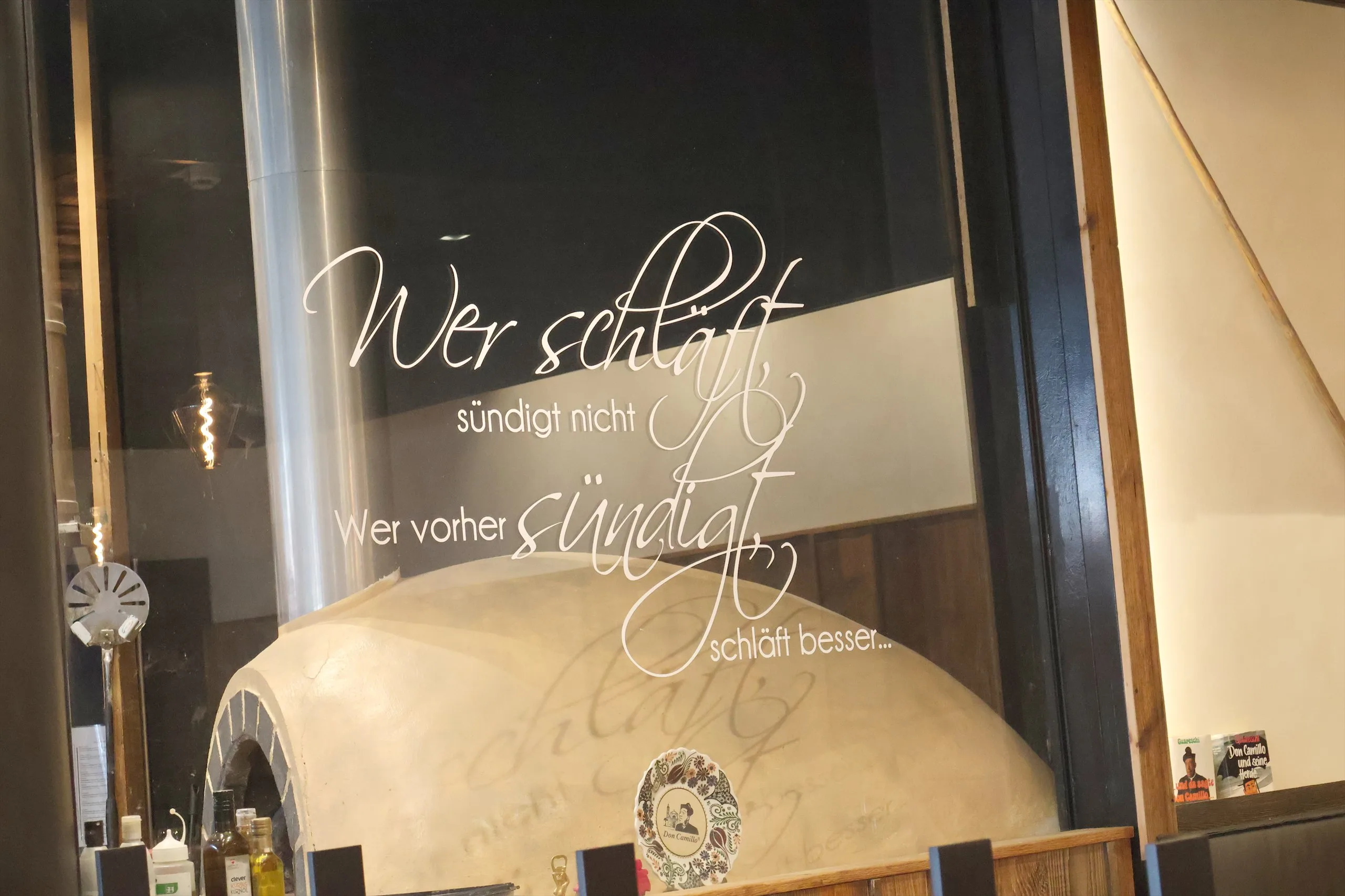 Gewölbter Pizzaofen im Lokal Im Stadioncenter, dahinter der Schriftzug „Wer schläft sündigt nicht“ auf der Glaswand